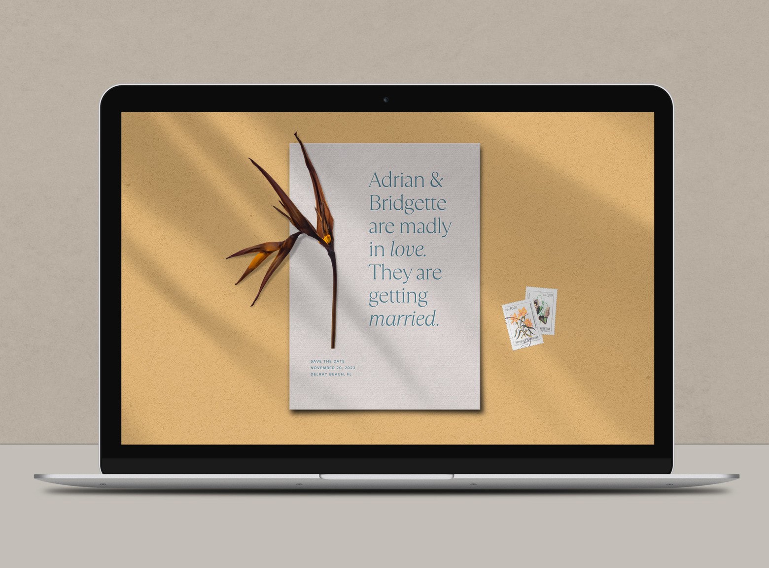 A laptop screen displaying a white virtual Save the Date with blue text, a yellow background and accented with a bird of paradise flower and stamps